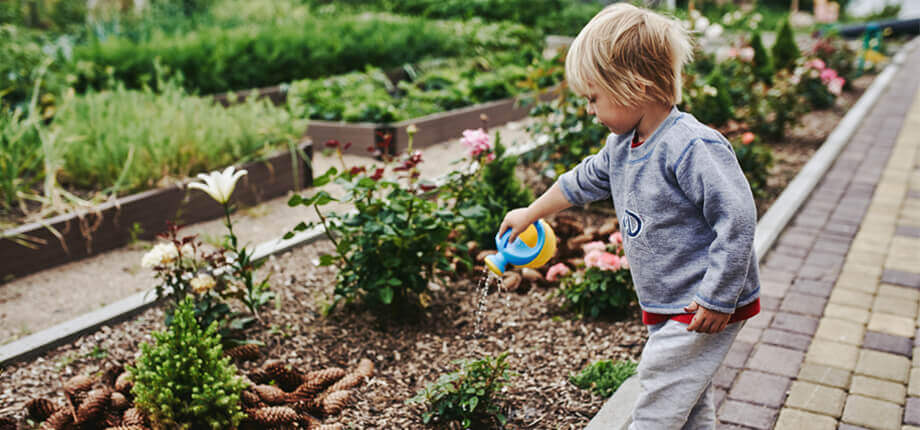 A boy with long blond hair is watering flower beds in the summer in the village